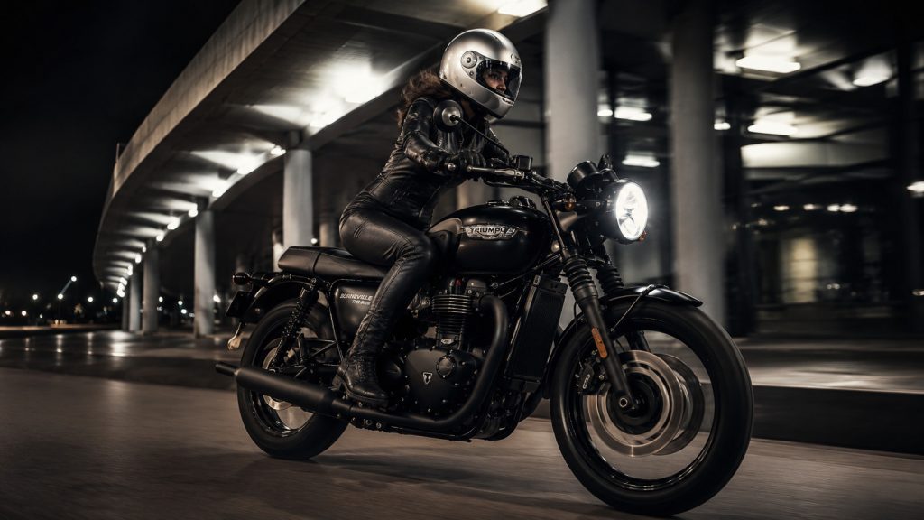 Female rider in a one-piece leather suit beside a black Triumph Bonneville T120-style motorcycle under urban night lights.